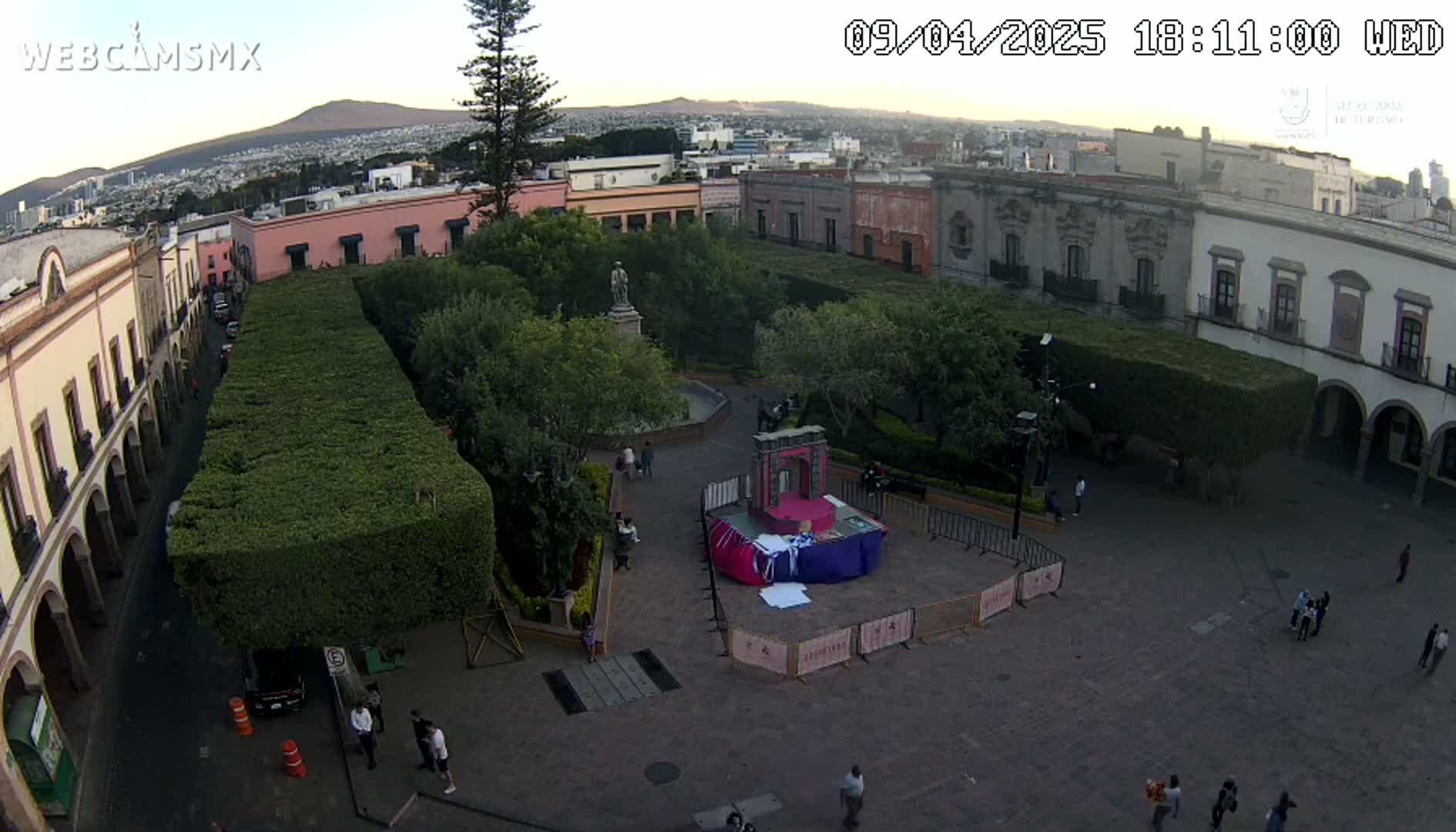 An aerial view shows a town square with a statue in the center, surrounded by buildings and neatly trimmed hedges under a clear evening sky.