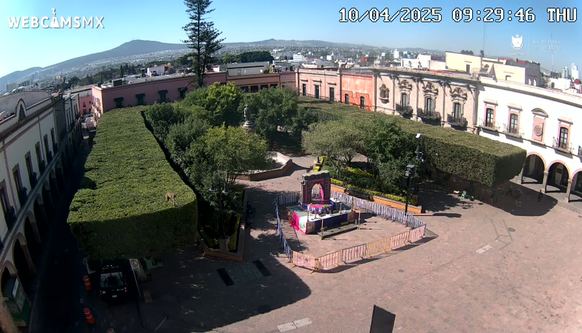 An aerial view shows a town square with trimmed hedges, colonial-style buildings, and a temporary altar in the center, under a clear, sunny sky.