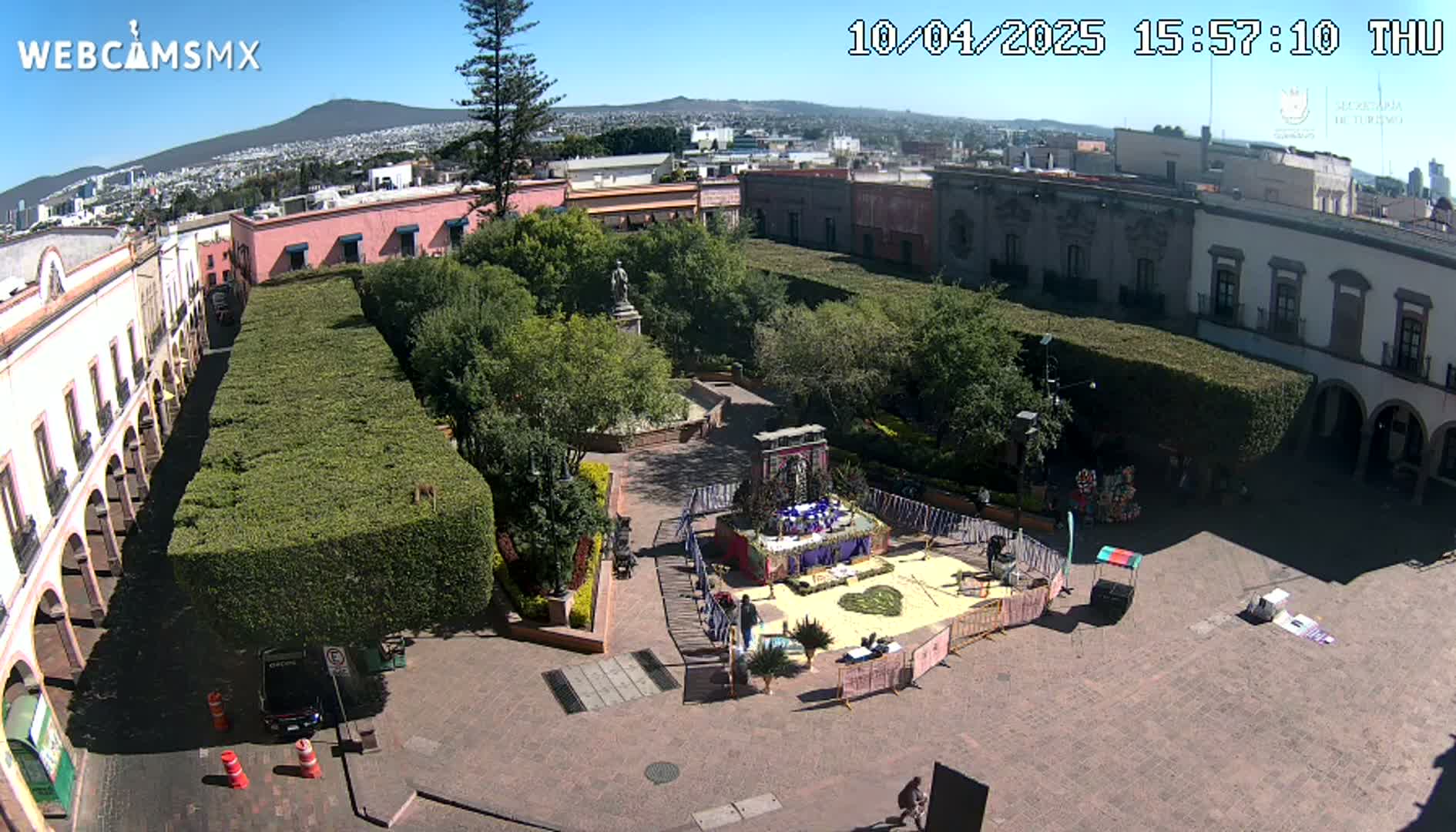 A sunny aerial view of a plaza with a central garden, surrounded by colonial-style buildings, and featuring a temporary altar-like structure in the plaza.