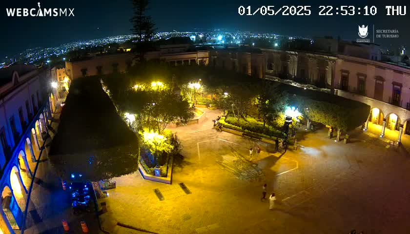 A nighttime, high-angle view of a brightly lit plaza surrounded by buildings, with a few people visible in the square and the city lights in the distance.