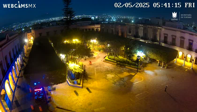 An illuminated plaza at night, surrounded by buildings, overlooks a city skyline under a clear, dark sky.