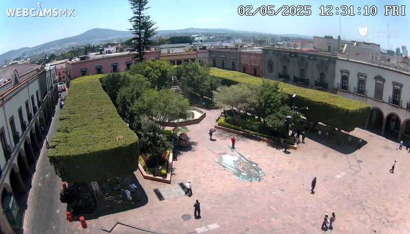 A sunny day reveals a town square with a central fountain, surrounded by neatly trimmed hedges, buildings, and a few people walking around.