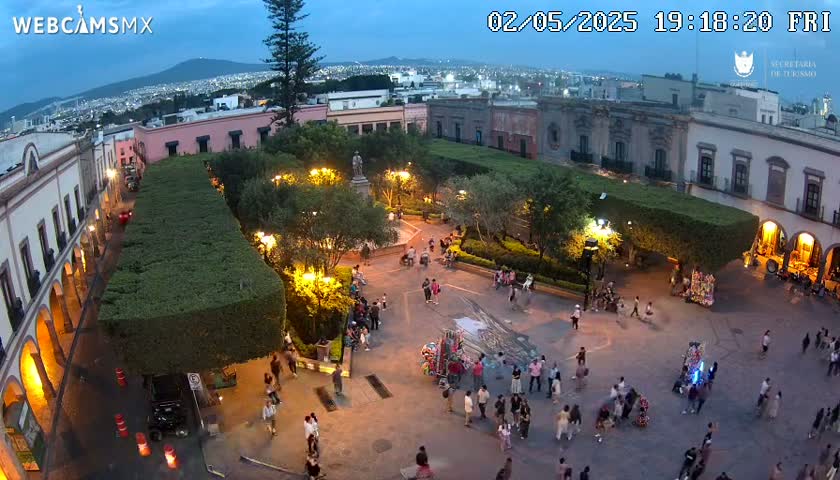An aerial, twilight view shows a town square with people gathered around a statue and vendors, surrounded by buildings and lush green hedges under a partly cloudy sky.