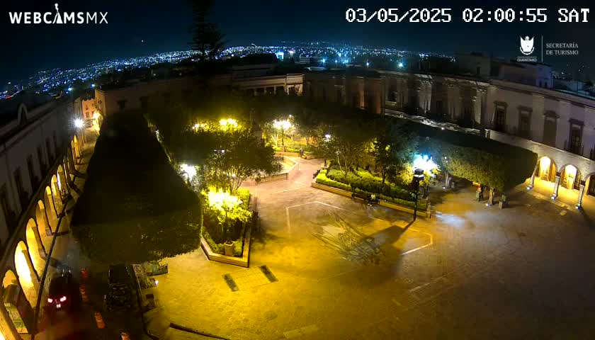A nighttime aerial view of a brightly lit plaza surrounded by buildings, with a city skyline visible in the distance.