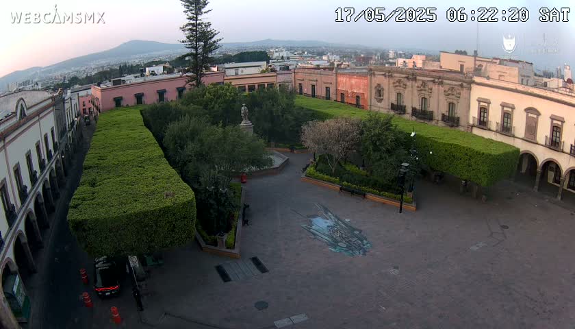 An aerial view of a plaza in a city, featuring colonial-style buildings surrounding a paved square with manicured hedges and a statue, under a clear, twilight sky.