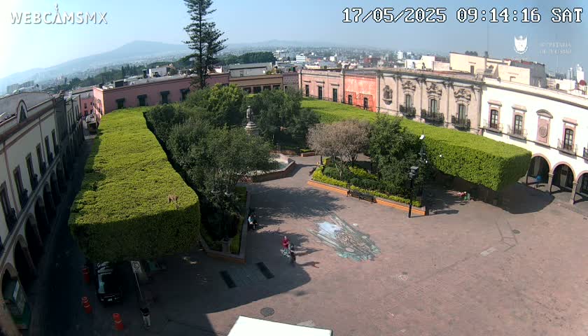 A sunny view of a plaza surrounded by colonial-style buildings, featuring a central park with manicured hedges and a small fountain, and a large street art piece on the ground.