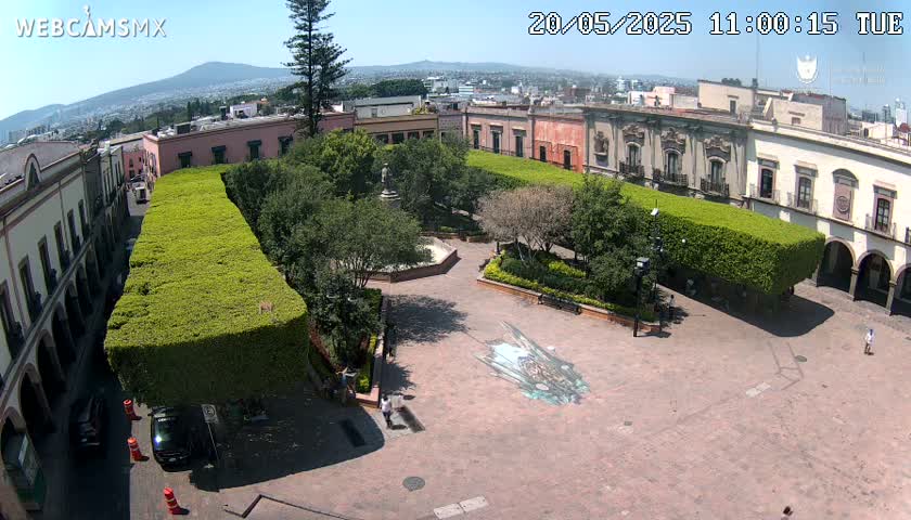A sunny aerial view of a town square with meticulously manicured hedges, colonial-style buildings, and a large, centrally located paved area featuring a ground mural.