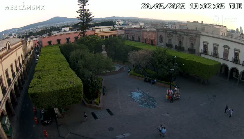 An aerial view shows a town square with colonial-style buildings, manicured hedges, a central fountain, and a few people walking around under a twilight sky.