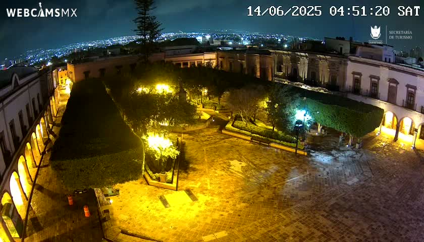 A nighttime view of a plaza, illuminated by yellow streetlights, shows a cobblestone square surrounded by colonial-style buildings and lush, trimmed greenery.