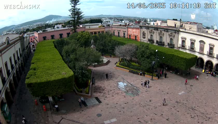A high-angle view shows a town square with a park in the center, surrounded by buildings, under a partly cloudy sky.