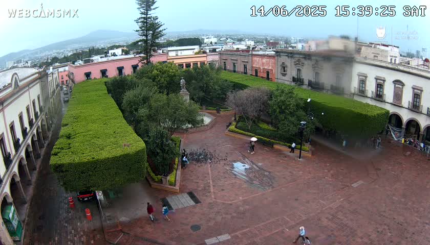 A high-angle view shows a town square with a park in the center, surrounded by buildings, under a partly cloudy sky.
