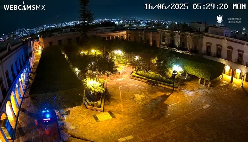 A nighttime aerial view of a town square, illuminated by streetlights, featuring buildings surrounding a paved plaza with landscaped areas.