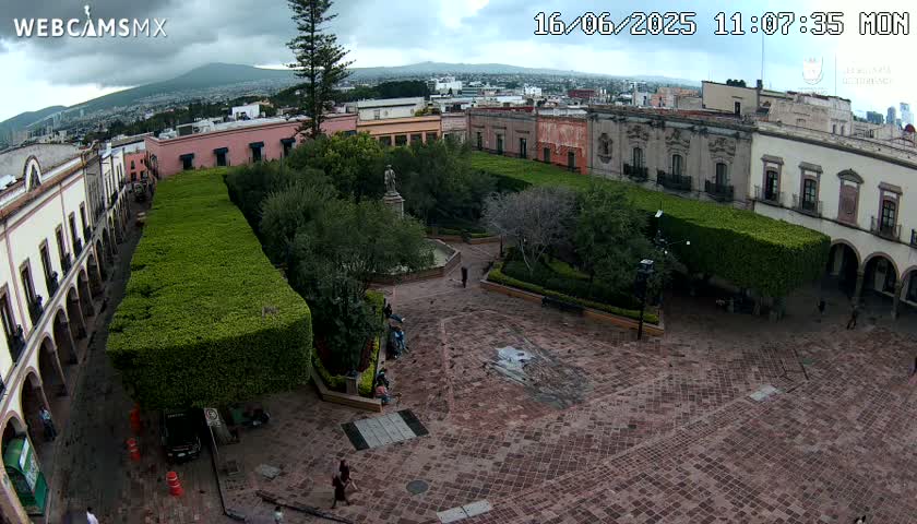 An aerial view of a town square with colonial-style buildings surrounding a central plaza featuring trimmed hedges, a fountain, and a statue, under a partly cloudy sky.