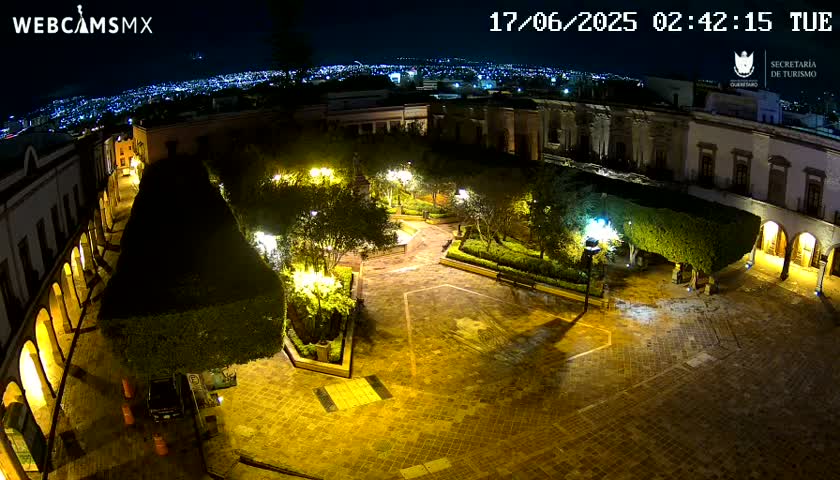 A nighttime, aerial view of a plaza with illuminated buildings and landscaping, overlooking a city in the distance under a clear sky.