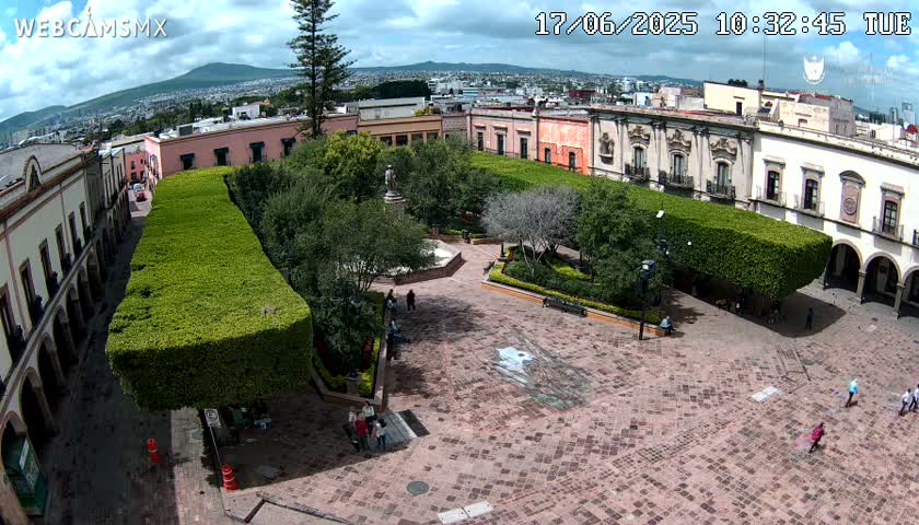 A high-angle view shows a town square with a paved plaza, surrounded by colonial-style buildings and neatly trimmed hedges under a partly cloudy sky.