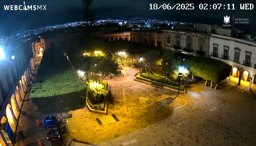 An illuminated plaza at night, surrounded by buildings and lush greenery, overlooks a city skyline under a dark sky.