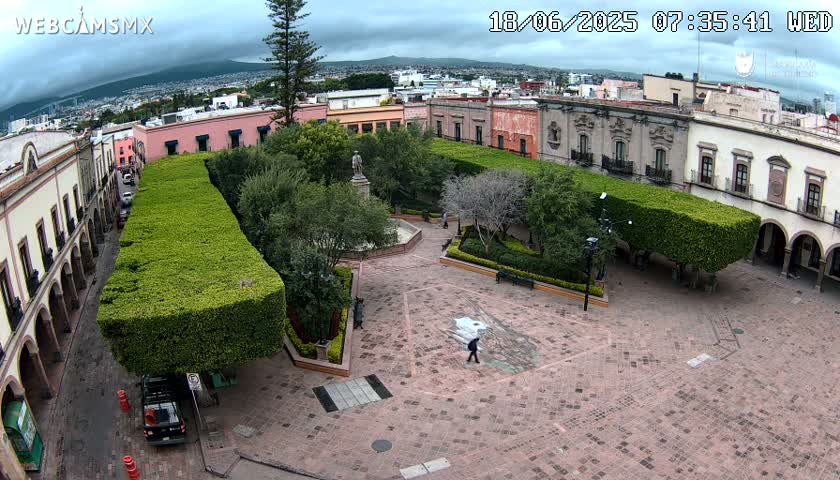 A paved plaza with a central garden and statue, surrounded by colonial-style buildings under an overcast sky.