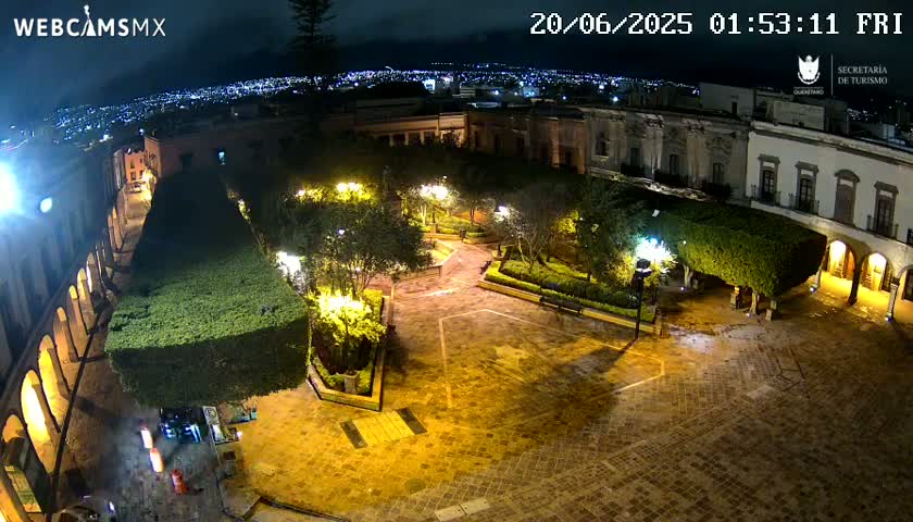 A nighttime view of a plaza, illuminated by lampposts, surrounded by low buildings and lush green shrubbery, with a city skyline visible in the distance under a partly cloudy sky.