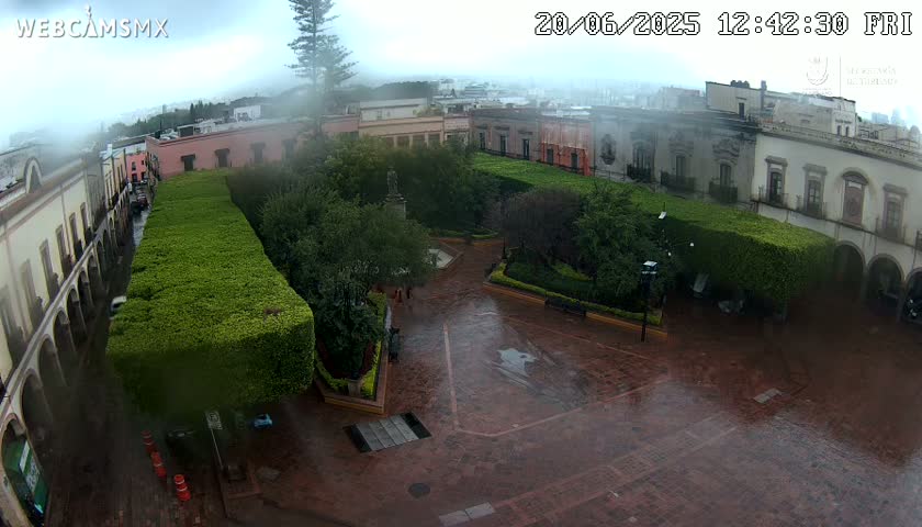 A rain-soaked town square, surrounded by colonial-style buildings and lush green hedges, is viewed from above.