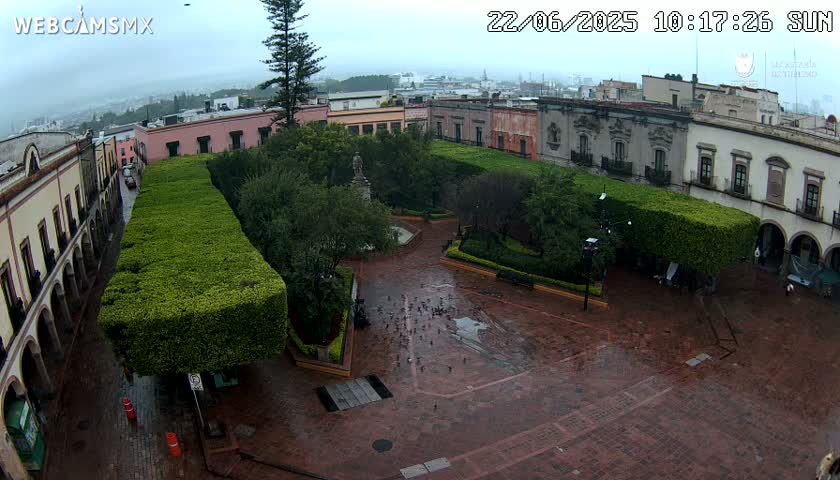 A brick plaza with a central park, surrounded by colonial-style buildings, is shown on an overcast day with light rain.