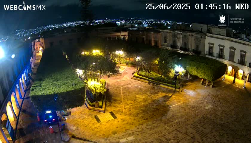 A nighttime aerial view of a brightly lit plaza surrounded by buildings, with shrubbery and trees in the plaza, and a city visible in the distance under a clear sky.