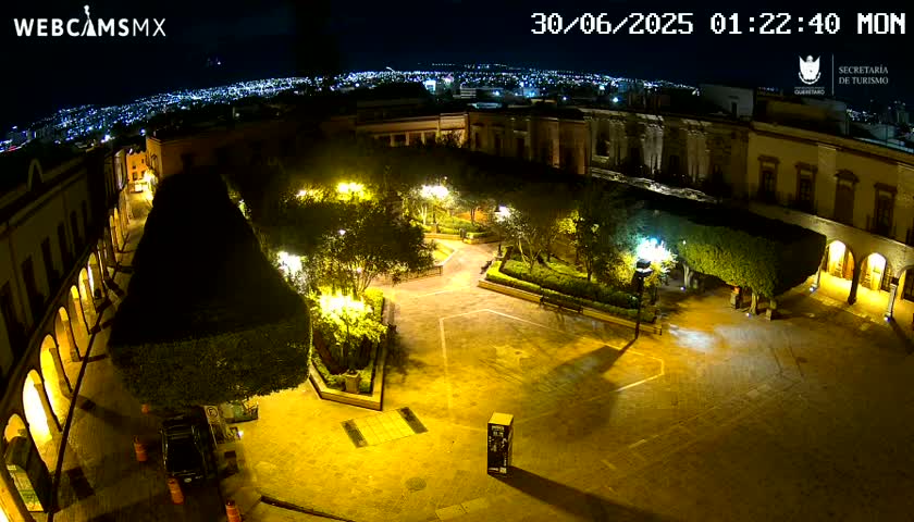 A nighttime, illuminated plaza with surrounding buildings and lush landscaping is visible, overlooking a city in the distance under a clear sky.