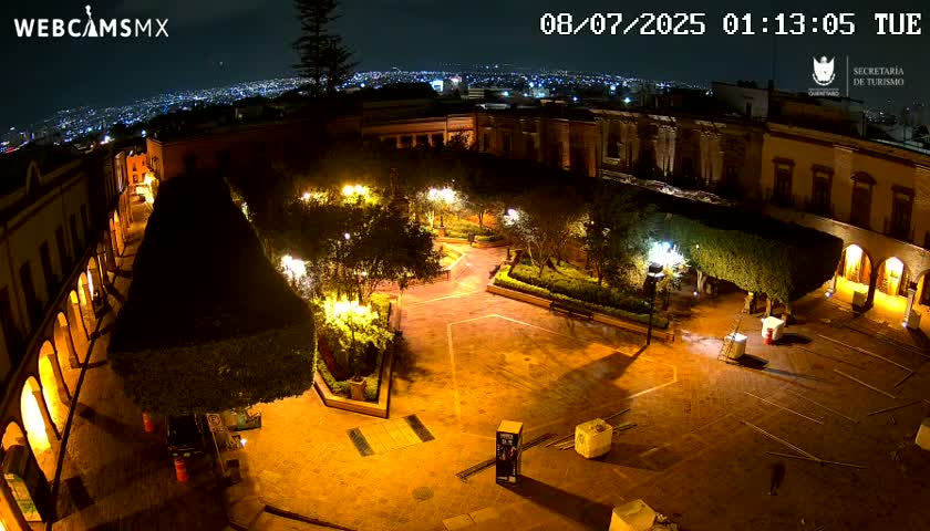 A nighttime aerial view of a plaza, illuminated by streetlights, surrounded by buildings, with a city skyline visible in the distance under a clear sky.