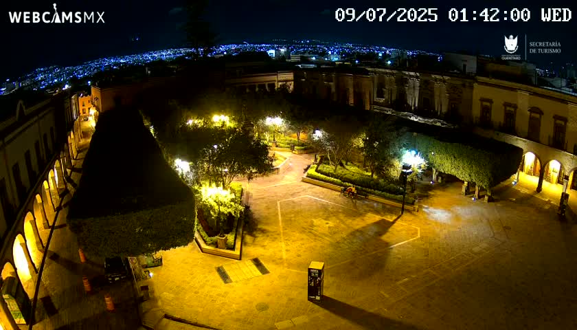 An illuminated plaza at night, surrounded by buildings, with a city skyline visible in the distance.