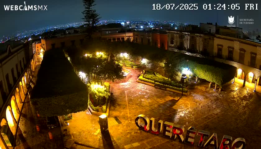 A nighttime view of a plaza with illuminated buildings, landscaping, and streetlights, showing wet pavement under a rainy sky.