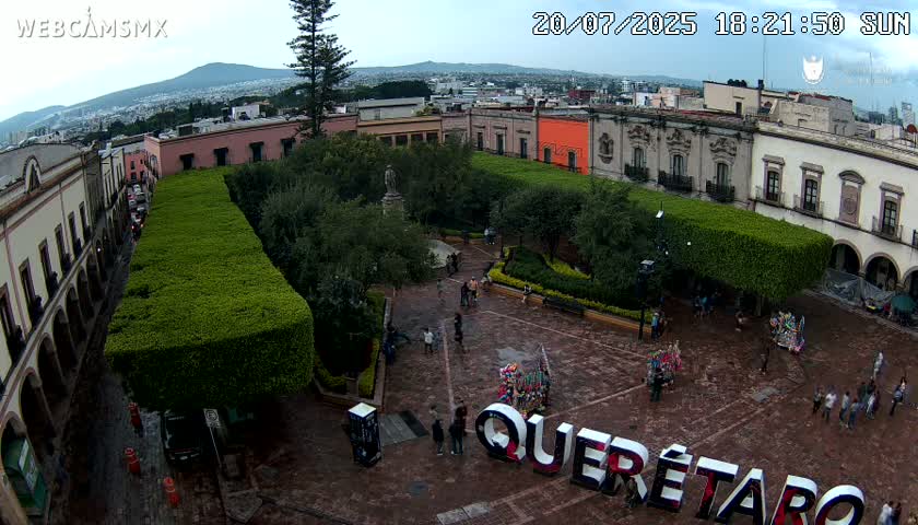 An aerial view of a town square with people walking around, surrounded by buildings and trimmed hedges under a partly cloudy sky.