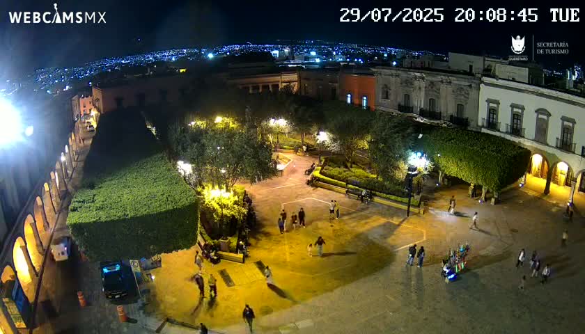 An aerial view of a town square with people walking around, surrounded by buildings and trimmed hedges under a partly cloudy sky.
