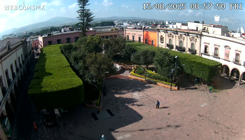 An aerial view of a town square with people walking around, surrounded by buildings and trimmed hedges under a partly cloudy sky.