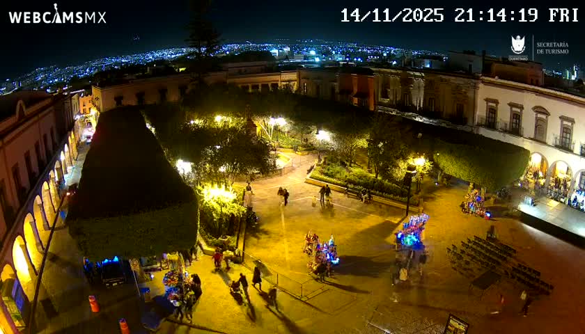 A vibrant city square, illuminated by warm yellow lights, bustles with people, vendors with glowing carts, and an event setup of chairs, all framed by historic buildings and a sprawling cityscape under a clear night sky.