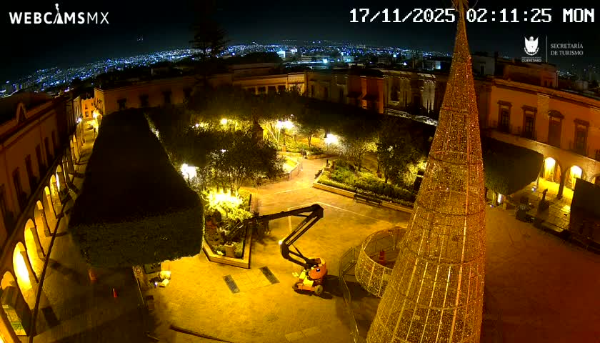 A brightly lit town square at night features a large, illuminated conical Christmas tree and a yellow boom lift, surrounded by traditional buildings, manicured trees, and a distant city skyline under a clear, dark sky.