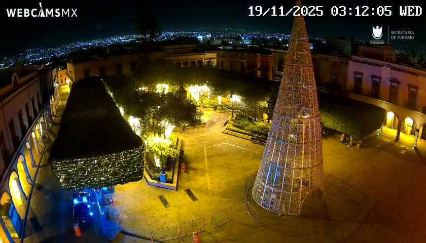 A brightly lit town square or plaza is visible at night under a clear sky, featuring a large, cone-shaped illuminated structure resembling a Christmas tree, other decorated trees and bushes, surrounding buildings with arches, and a distant city skyline glowing in the background.