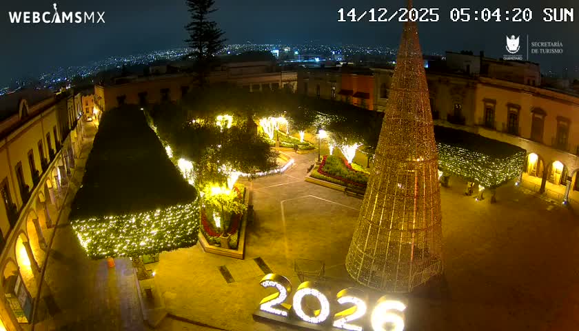 A brightly lit town square is seen at night, featuring a large, illuminated Christmas tree-like structure, surrounded by buildings and a distant cityscape under clear, dark skies.