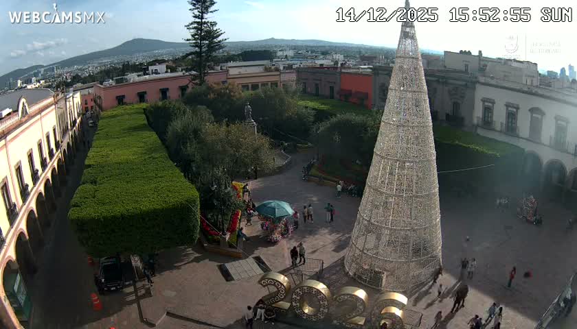 A brightly lit town square is seen at night, featuring a large, illuminated Christmas tree-like structure, surrounded by buildings and a distant cityscape under clear, dark skies.