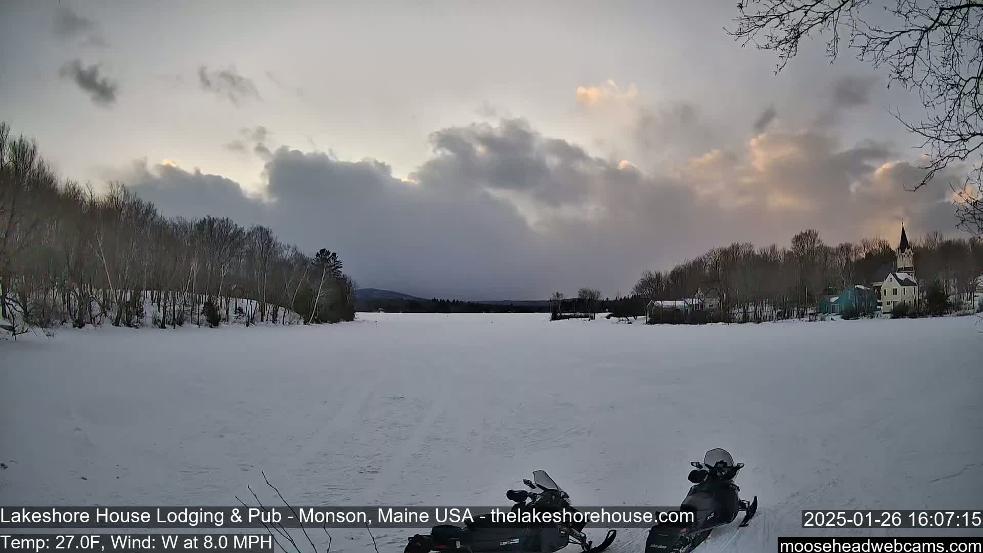 A snow-covered frozen lake is seen, with a village and trees in the background under a cloudy, dusky sky.