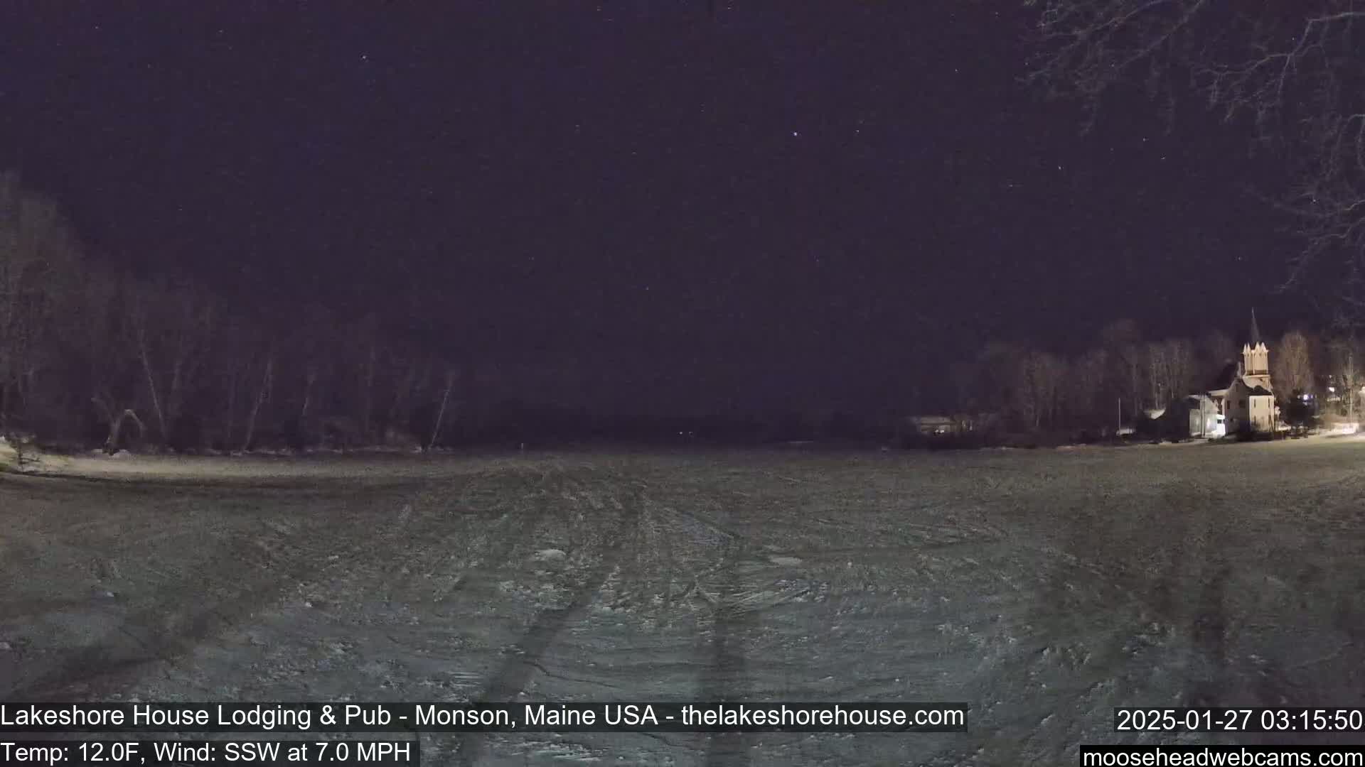 Under a clear, starry night sky, a snow-covered landscape stretches out before a dark treeline and a building, with a south-southwest wind blowing at 7 mph and a temperature of 12 degrees Fahrenheit.