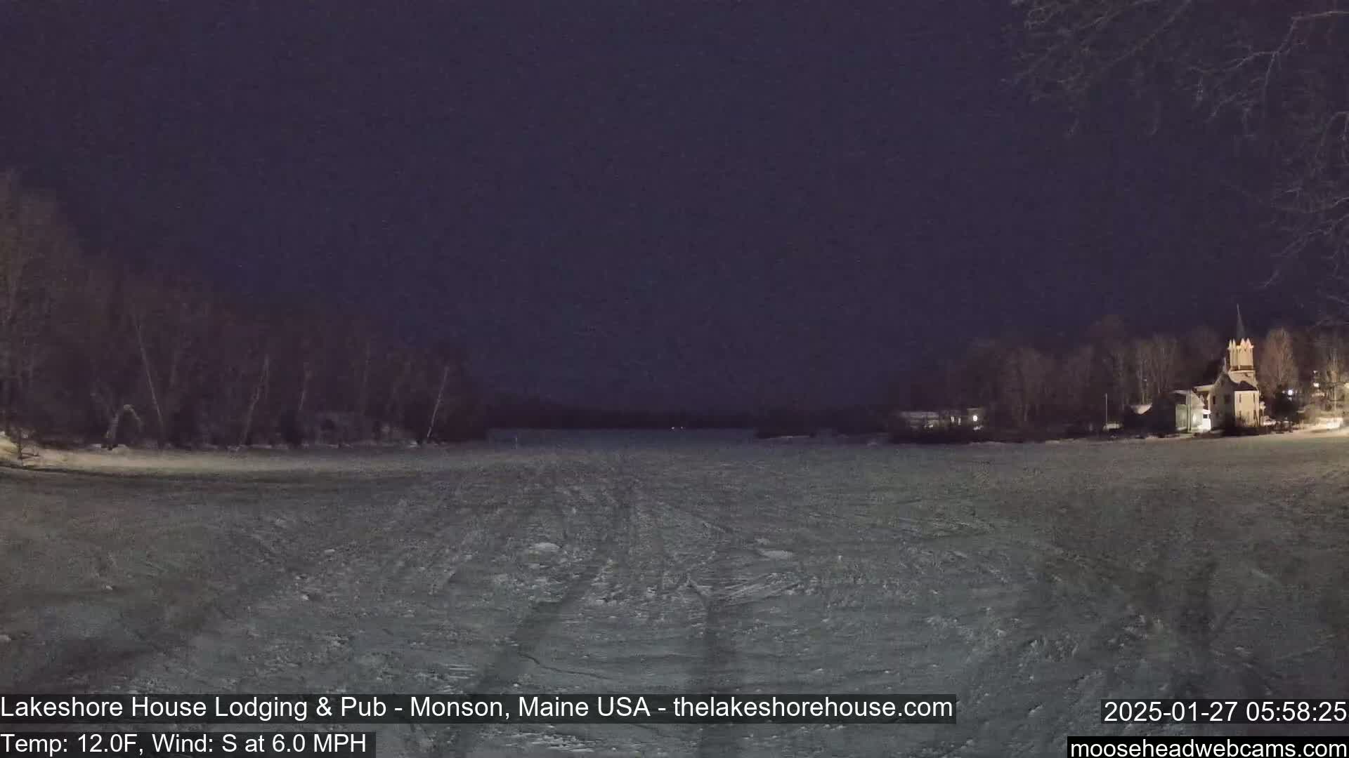 A nighttime view of a frozen lake under a dark sky, with a building visible on the far shore and trees lining the lake's edge.