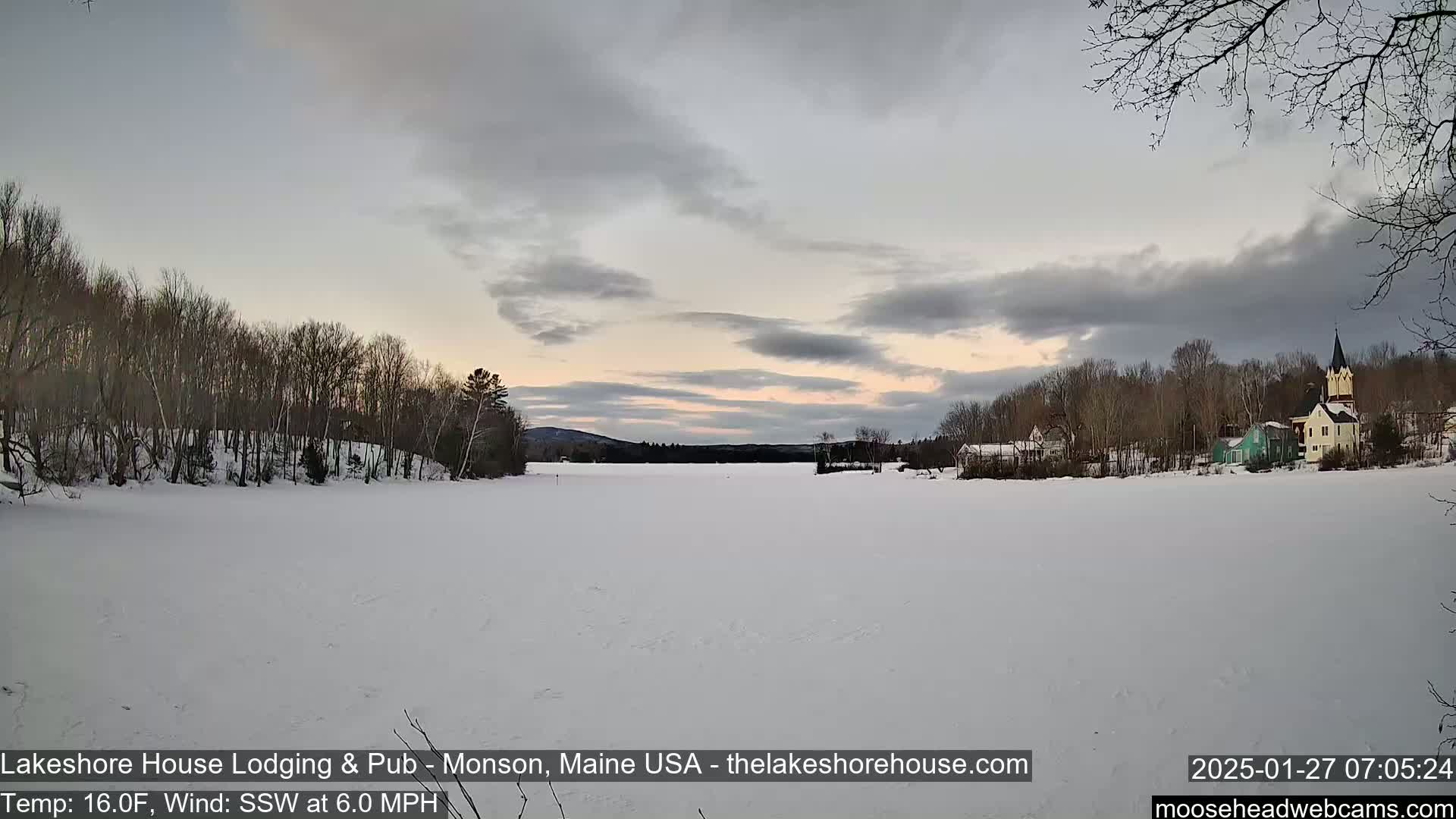 A frozen lake stretches out before a small village under a mostly cloudy, pale sky at sunset.