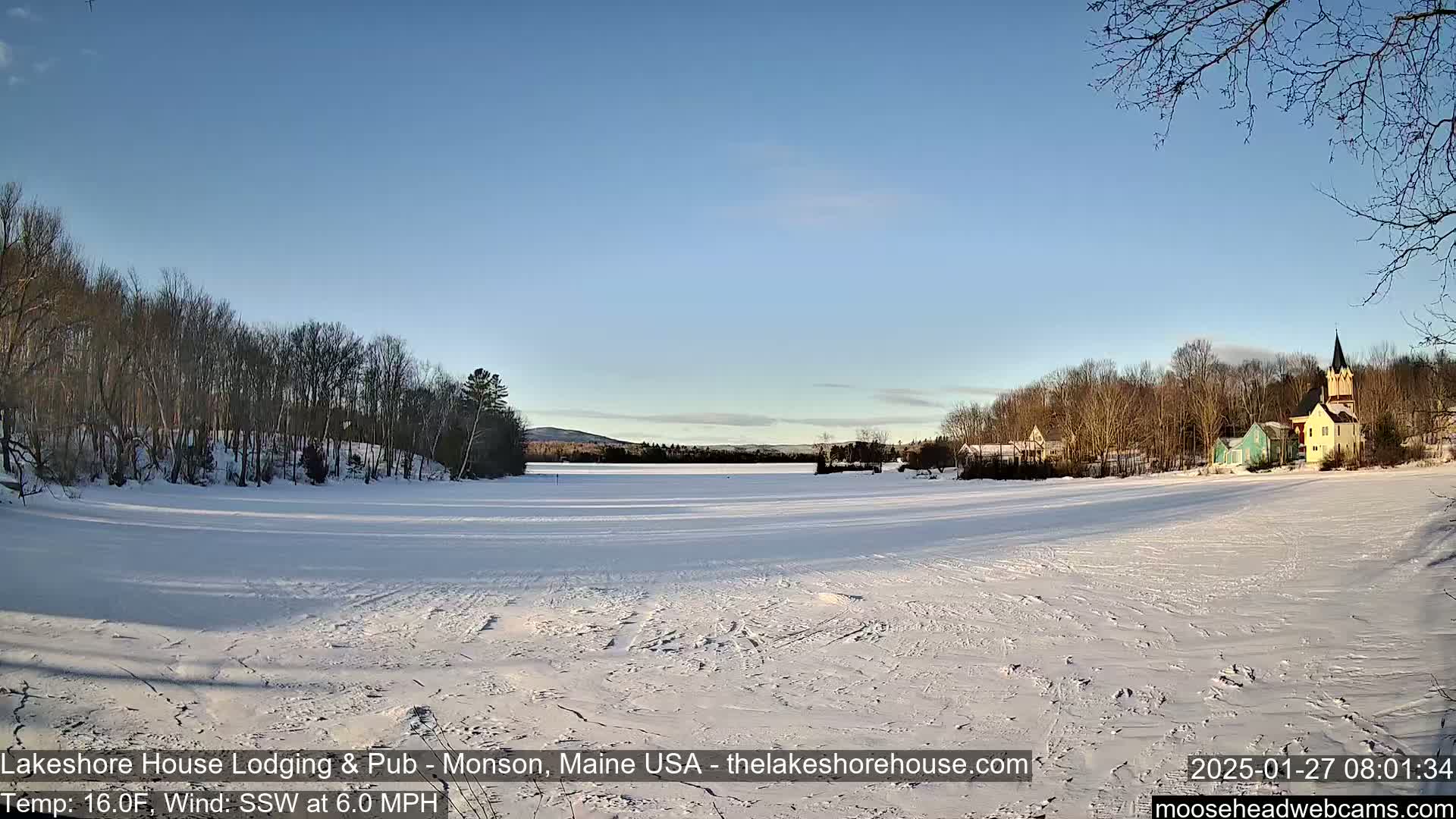 A frozen lake, covered in snow with tracks, is situated in front of a small village nestled among leafless trees under a clear blue sky.
