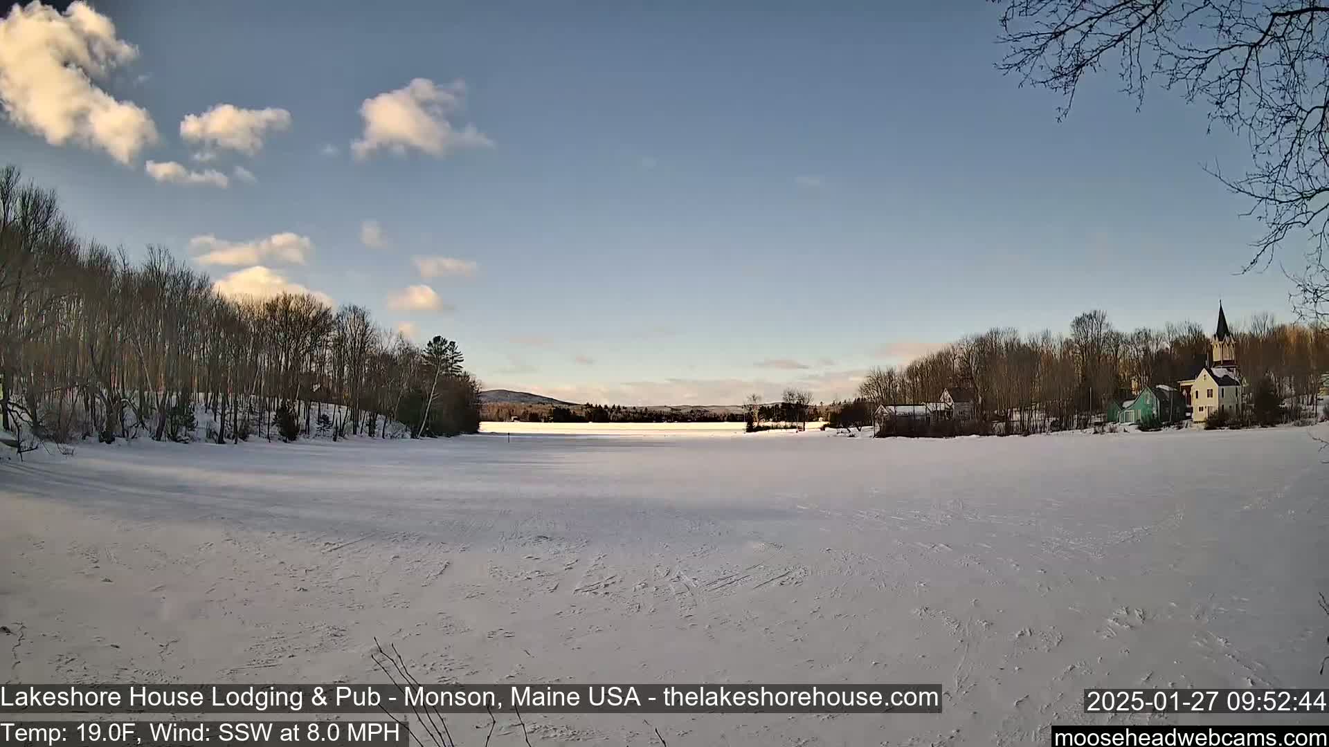 A frozen lake stretches out before a snow-covered shoreline dotted with buildings and trees under a partly cloudy sky.