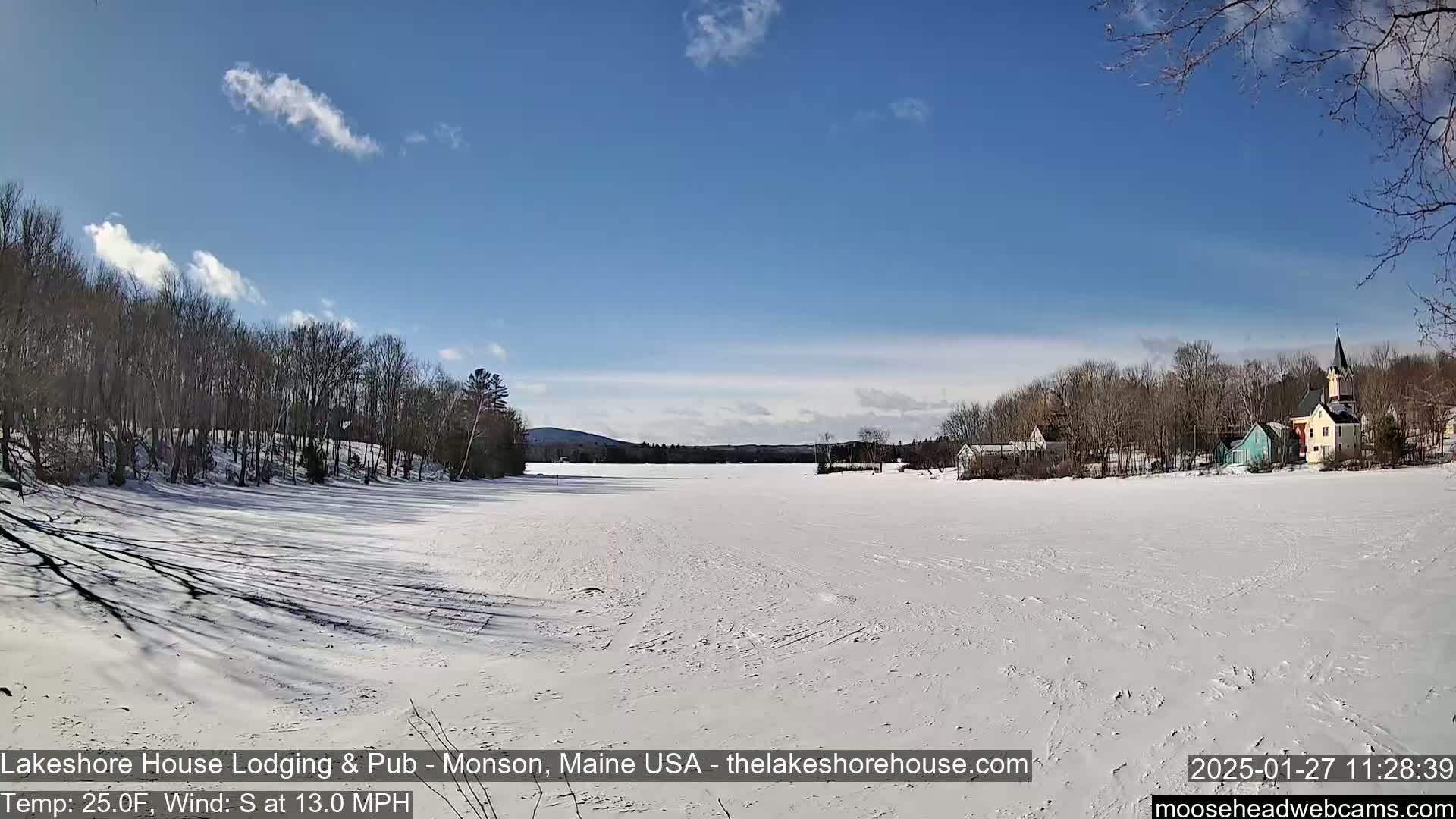 A snow-covered frozen lake stretches out before a line of trees and houses under a mostly sunny sky.