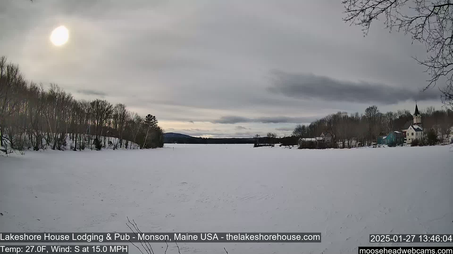 A frozen lake under a mostly cloudy sky with a bright sun, surrounded by snow-covered trees and a small town in the distance.