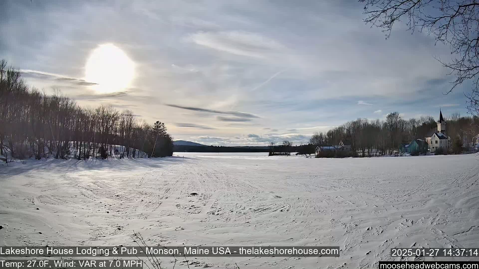 A snow-covered frozen lake stretches towards a village with trees and buildings under a bright, sunny sky with thin clouds.