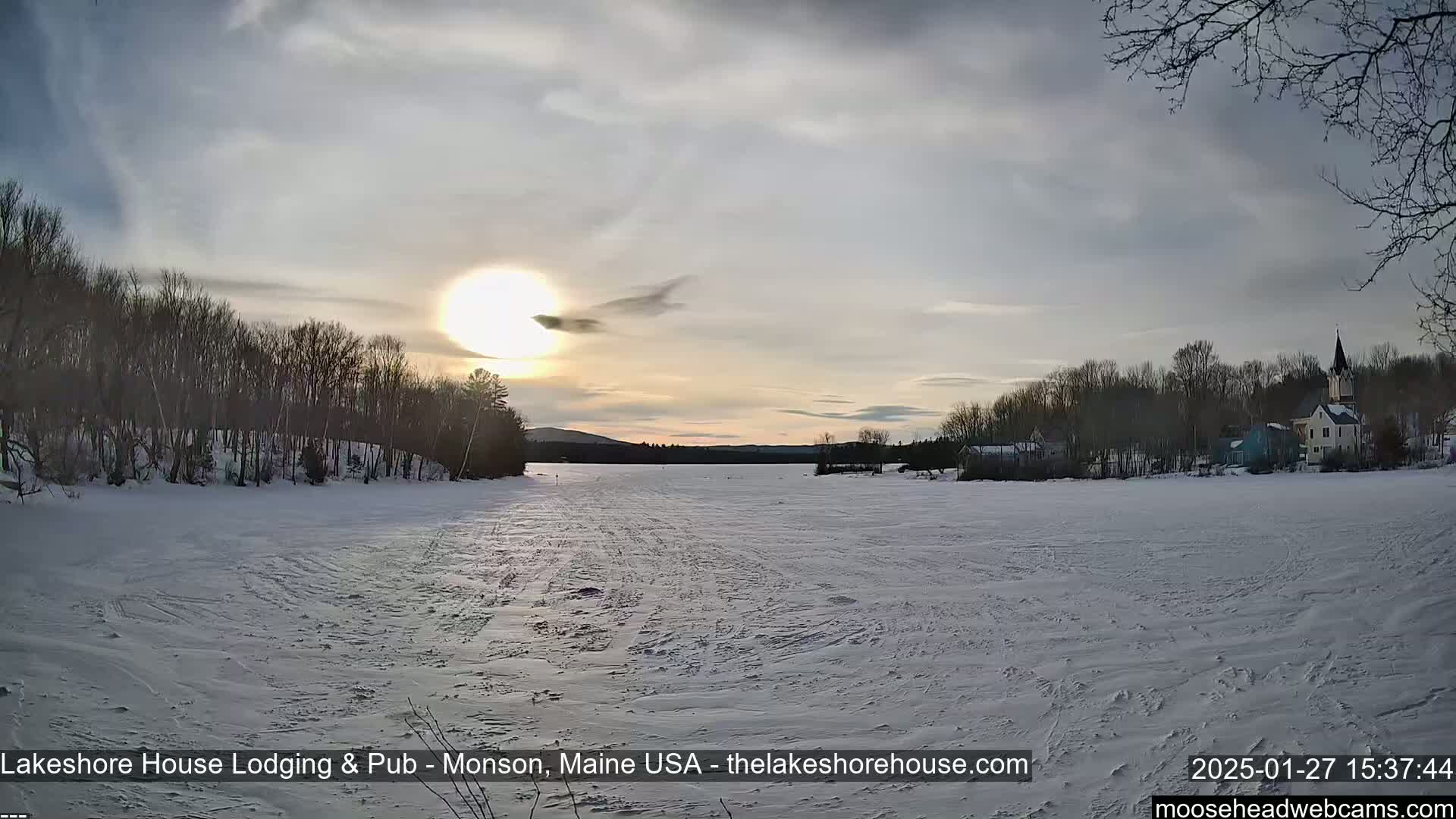 A frozen lake stretches out under a partly cloudy sunset, with snow-covered trees and buildings lining the far shore.