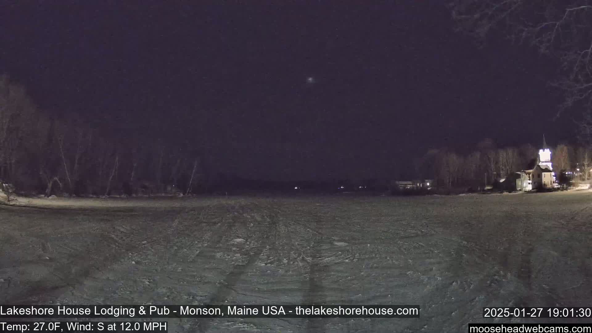 A snow-covered, frozen lake at night is seen under a dark sky, with trees and buildings visible in the distance.