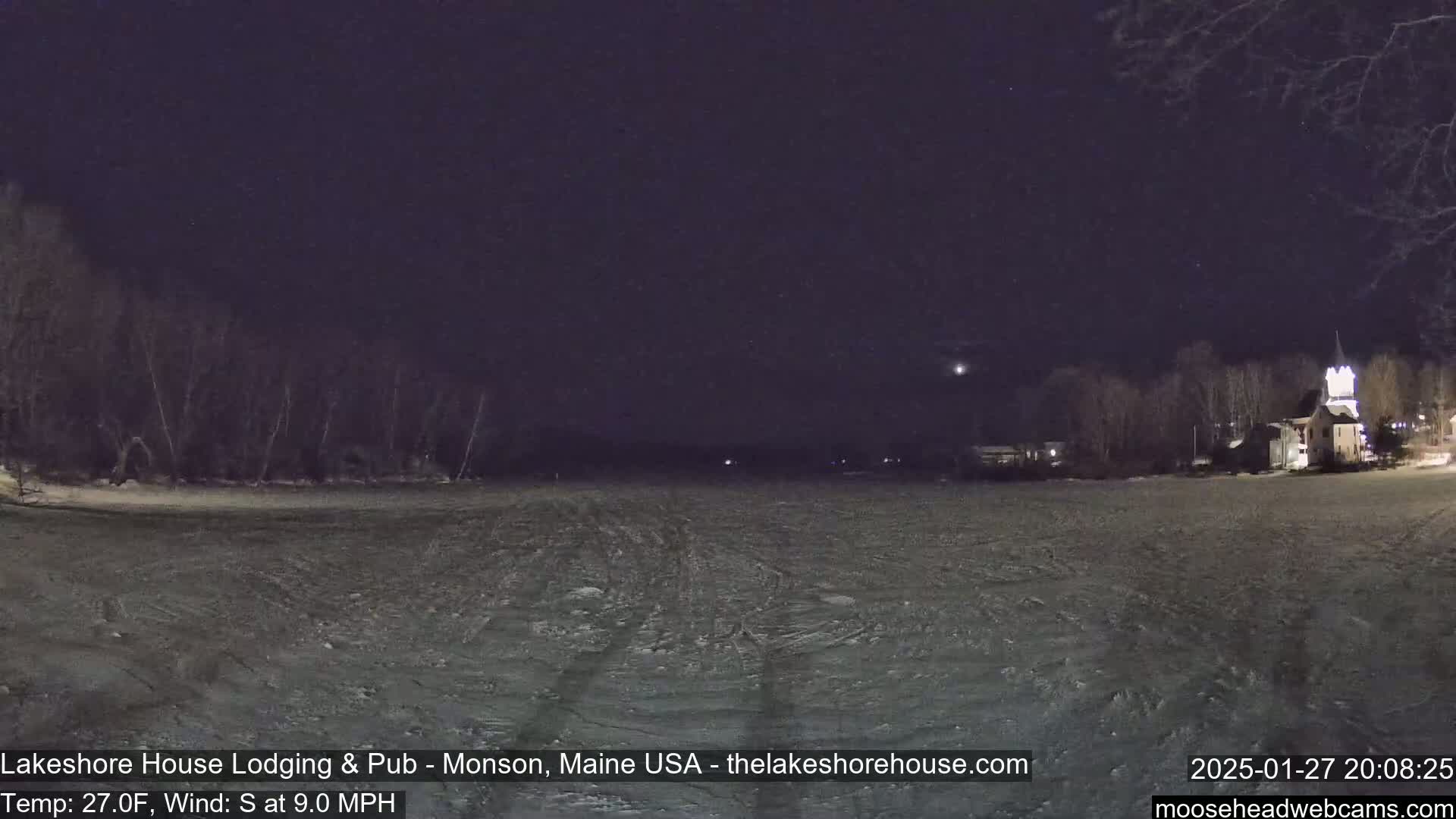A snow-covered, dark expanse stretches out under a night sky, with a dimly lit town and trees visible in the distance, a slight south wind blowing at 9 mph and a temperature of 27°F.