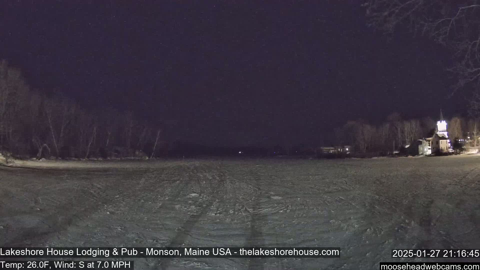 A frozen lake at night, under a dark sky, with trees and a building visible in the distance, and a south wind blowing at 7 mph.